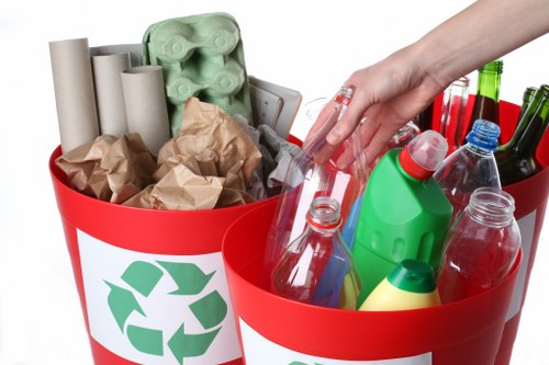 A close-up image of two red recycling bins with green recycling symbols on their sides, filled with various recyclable waste items. The left bin contains several rolled-up brown and beige paper and cardboard, along with a few crumpled paper bags. The right bin holds empty plastic bottles of different sizes and colors, including transparent, green, and blue, some with caps still attached, and a few plastic containers. A person's hand is reaching into the right bin to place or retrieve an item. The scene emphasizes household recycling and waste separation, with the bins placed against a plain white background. The image showcases common recyclable materials such as paper and plastic, relevant to waste management services in Twickenham, emphasizing environmentally conscious rubbish removal for domestic or office environments in the local area.