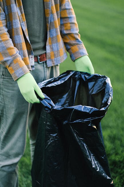 A collection of overflowing rubbish bins and scattered waste bags, cardboard boxes, paper, and plastic packaging are piled on the asphalt pavement in front of a commercial building. The large grey bin