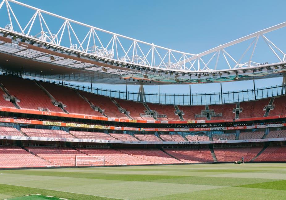 A clear view of an empty football stadium during daylight hours, with a well-maintained green pitch in the foreground. The seating area features red and grey seats arranged in multiple tiers, with som